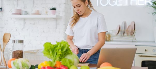Woman in white t-shirt preparing fresh vegetables in a bright kitchen with a laptop.